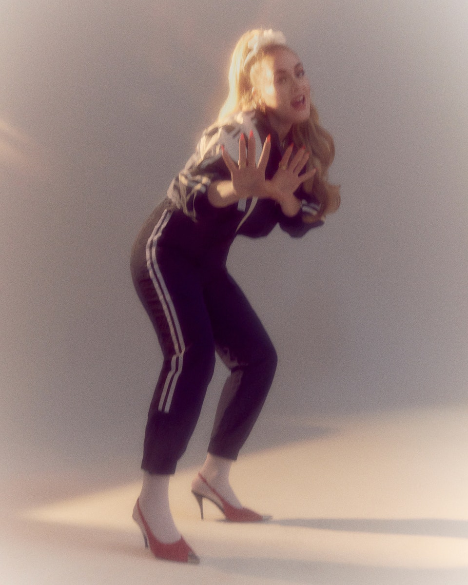 Adele is wearing a Balenciaga tracksuit and earrings by Moya. Her hair is tied up in a scrunchie, and she is wearing pink Saint Laurent high heels with while socks. Shot by Charlotte Wales for The Face Magazine.